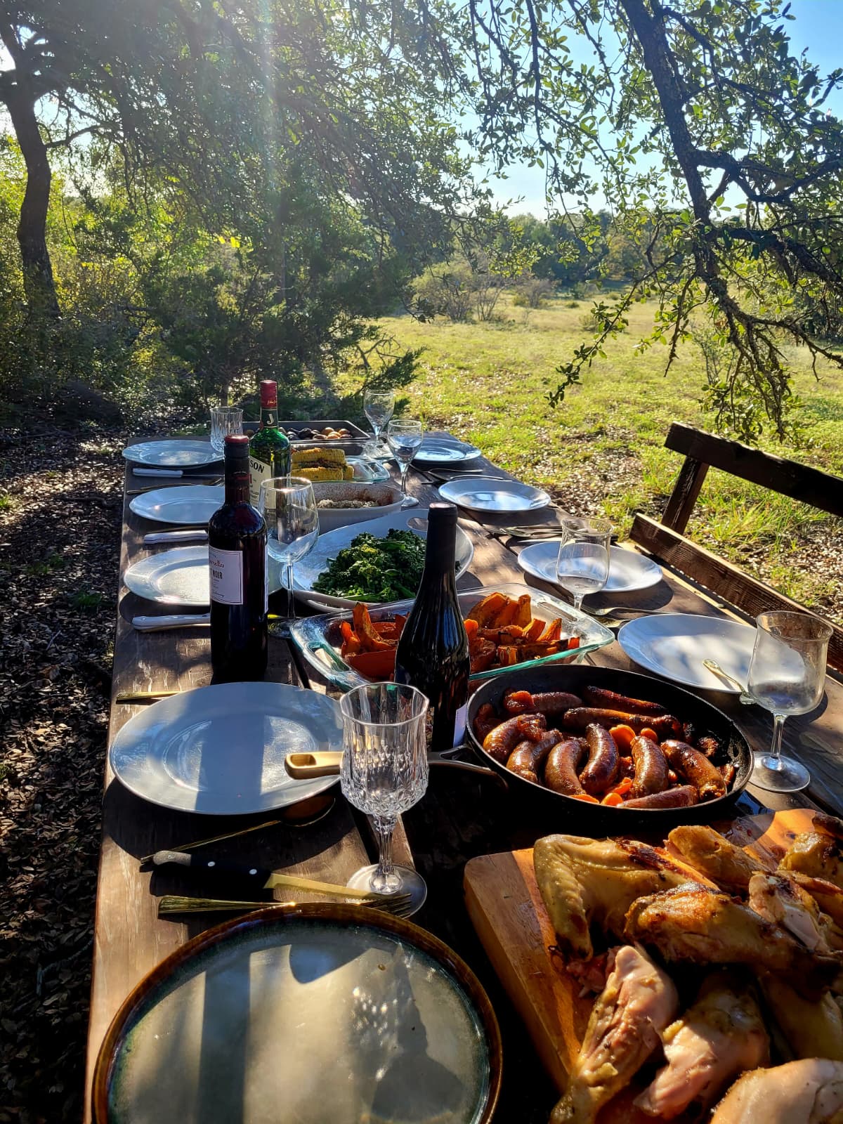 A long table laid with roast chicken, sausages and sides under the oak trees